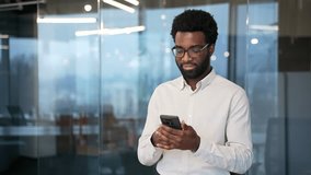 African american businessman is using browsing mobile phone standing in a modern glass business office. Worker is chatting online with a client, working in app or reading writing message on smartphone - Powered by Shutterstock - Get 15% off with code: PIKWIZARD15
