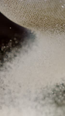 Extreme close up shot of a cook sifting flour or powdered sugar through a metal sieve for a recipe, preparing a cake batter or bread dough in a professional kitchen against a dark background