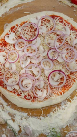 Professional chef preparing an authentic italian pizza, sprinkling seasoning over the tomato sauce, cheese, onions, and mushrooms. The wooden board is surrounded by fresh, colorful ingredients