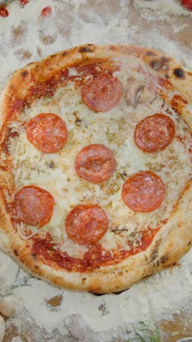 Top-down view of people taking slices of freshly baked pepperoni pizza from a wooden board, surrounded by fresh ingredients like tomatoes, peppers, and onions on a rustic table