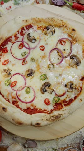 Top-down view of a freshly baked vegetarian pizza with mozzarella, mushrooms, and onions rotating on a wooden board, surrounded by colorful vegetables, spices, and scattered flour on the table