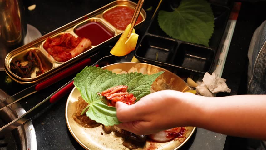 Hand prepares Korean BBQ wrap with perilla leaf, radish, garlic, and beef under warm lighting