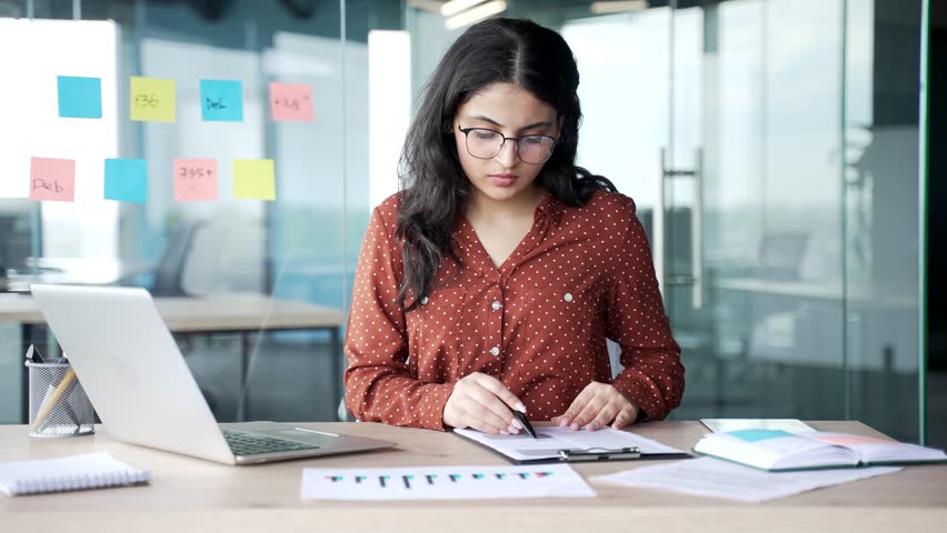 Confident young businesswoman signing documents sitting at a desk at workplace in a business office. Female executive looks through the folder with documentation, checks and signs contract with a pen