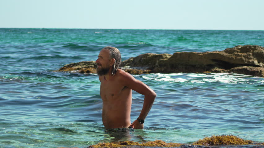 Man beard sea, standing in turquoise clear water by rocky shore during summer vacation, enjoying healthy lifestyle
