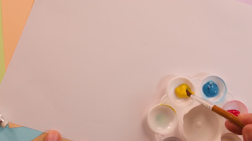 Hand mixes yellow and blue paint on palette, creating green, under soft lighting, overhead view