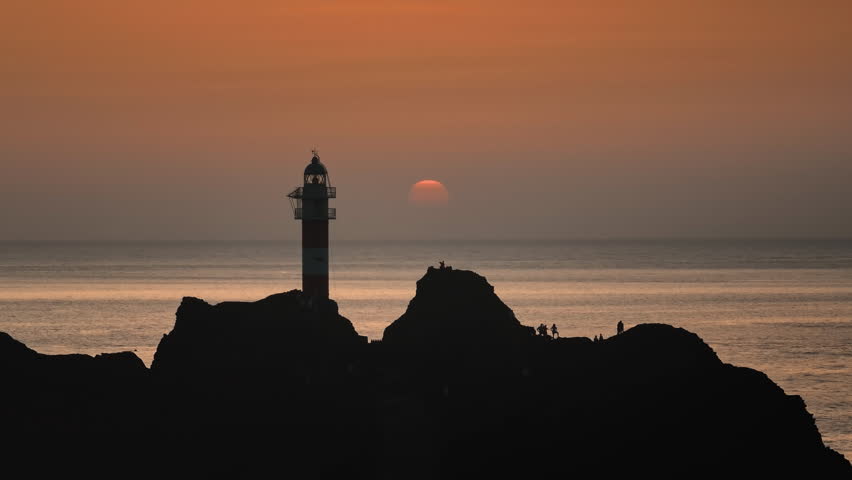 Punta de Teno lighthouse silhouette in Tenerife, with Atlantic Ocean view under colorful evening sky. Drone flight footage panorama. Canary Islands: Tourists enjoy vibrant orange sunset landscape