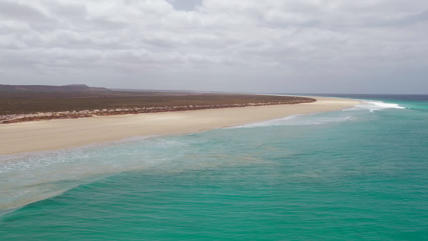 Aerial footage of Santa Monica Beach,showcasing endless white sand, turquoise waves, and a pristine,background volcanic mountain and desert,Boa Vista, Cape Verde.