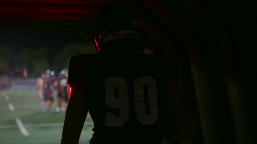 Silhouetted american football player with number 90 walking from a dark tunnel onto a brightly lit stadium field at night, ready for the big game with the team waiting in the background