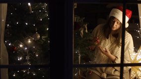 Happy woman in santa hat and cozy sweater decorating stylish christmas tree with modern baubles, view from window. Merry Christmas and Happy Holidays! Family xmas tradition video - Powered by Shutterstock - Get 15% off with code: PIKWIZARD15