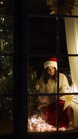 Happy woman in santa hat and cozy sweater putting stylish christmas present from santa bag under xmas tree with lights, view through window. Merry Christmas! Magical eve vertical footage