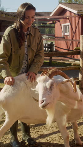 Vertical shot of young female animal caretaker smiling at camera while petting goat inside wooden paddock on farmyard, tending livestock on farm, slow motion