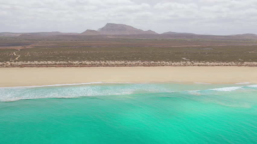Aerial footage of Santa Monica Beach,showcasing endless white sand, turquoise waves, and a pristine,background volcanic mountain and desert,Boa Vista, Cape Verde.