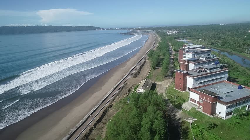 Aerial view of wide sandy beach with ocean waves rolling in, bordered by a green forested area and shoreline protection infrastructure along the coastline