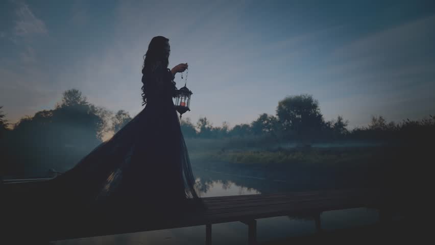 Fairy tale silhouette gothic woman hand hold lantern lamp walking across bridge over river dense fog mist night nature dark dramatic blue sky. long black dress back rear view. Fantasy scared girl.