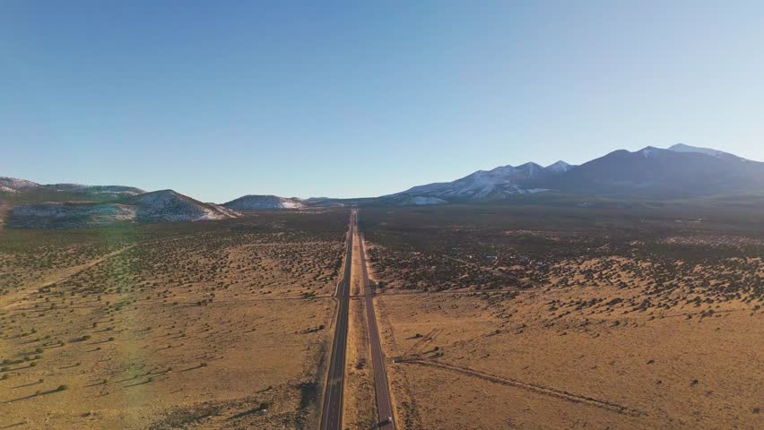 Aerial view of a road cutting through the landscape toward snow-capped mountains of the San Francisco peaks under a clear sky, Flagstaff, Arizona, United States.