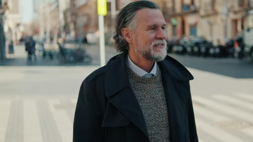 A mature man with a beard and long gray hair smiles as he walks down a city street. He wears a black trench coat and a gray sweater.