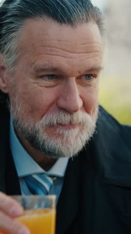 Vertical video, A mature businessman enjoys a glass of orange juice while checking his phone outdoors. He takes a moment to savor his drink and engage with his mobile device.