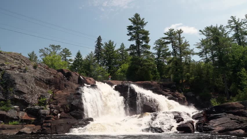 A small figure of a person standing at the top of a large waterfall surrounded by rocks and trees. Seen from a boat in the water in High Falls Park