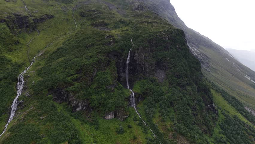 An aerial view of a Nordic mountain landscape with steep slopes, cascading waterfalls, a glacial lake, and a winding road through a misty green valley - Powered by Shutterstock - Get 15% off with code: PIKWIZARD15