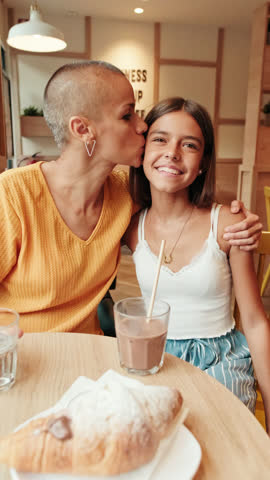 Happy young girl smiling at the camera in a cafe. She is wearing a white top and a gold necklace. She has long brown hair and is sitting at a table.
