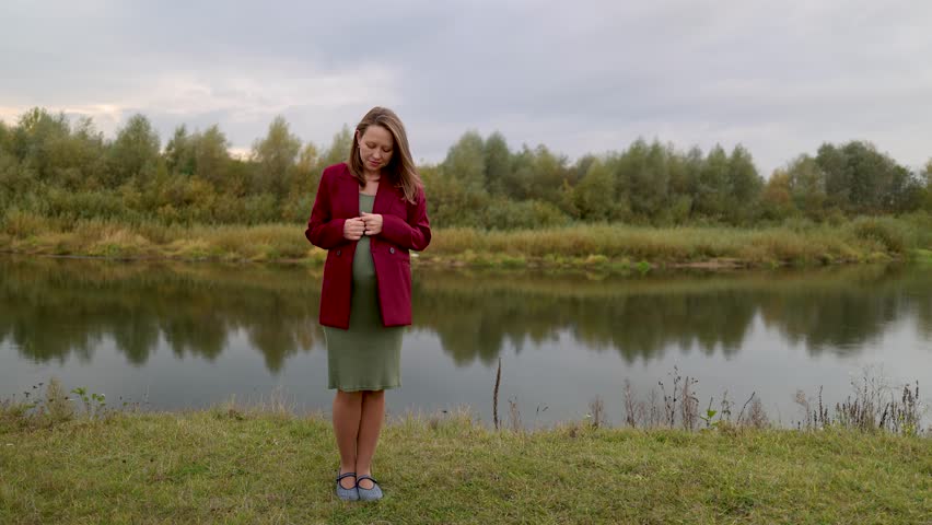 A pregnant woman in a green dress on the riverbank. A girl in a green dress. The expectant mother gently strokes her belly with her baby. Cloudy weather. Autumn park.