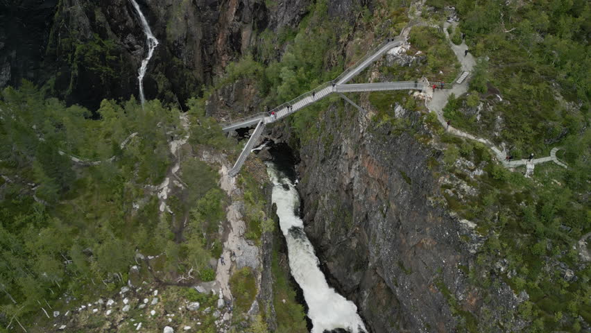 Drone shot of the bridge at the tourist attraction Vøringsfossen Waterfall in Norway. People walk across the bridge and cross a steep valley through which the water flows to the waterfall.
