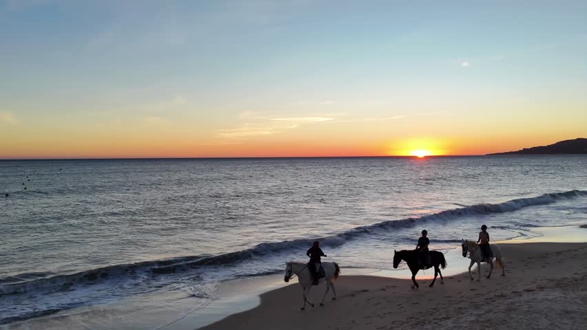 An aerial view of three people on horseback ride along a sandy beach at sunset in Costa de la Luz, Spain