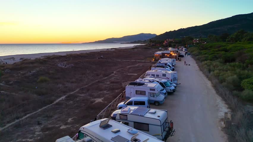 An Aerial view of Line of camper vans along coastal dunes with green forest, calm ocean, road and hills at sunset in Tarifa, Spain