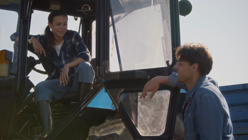 Young female farmer in work clothes sitting in tractor cabin while chatting with cheerful man during crop harvest season on farmland