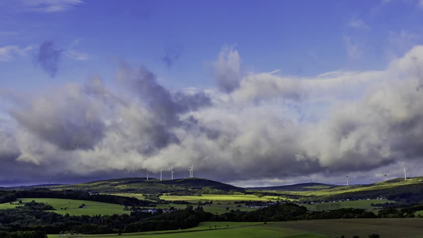 A timelapse of dramatic landscape of rolling green hills with distant wind turbines under a stormy, cloudy sky. A faint rainbow is visible