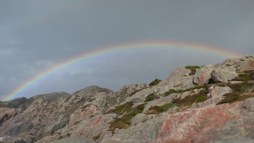 A vibrant rainbow stretches across the sky, creating a stunning contrast against rocky cliffs and patches of green moss under a dramatic sunset sky.