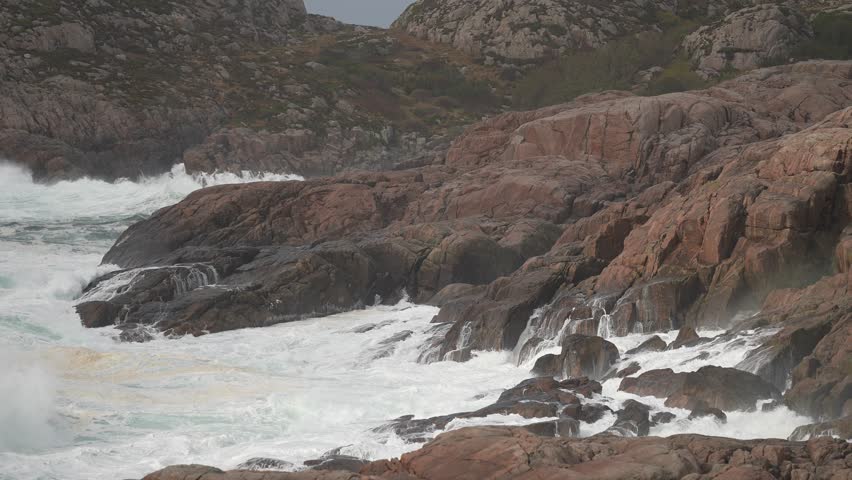 Powerful ocean waves crash against rugged rocks along a wild coastline. The scene captures the raw beauty of nature during stormy weather, creating dramatic splashes and mist.