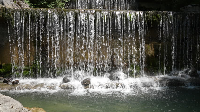 A slow-motion shot captures water cascading down a small, tiered fall surrounded by lush greenery near Walensee, Schweiz. Concept: Contemplative flow and engineered nature.