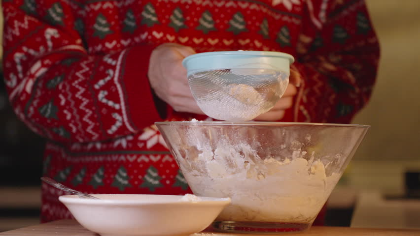 Woman in Red Sweater Sifting Flour into Mixing Bowl While Baking in Festive Kitchen During Evening Christmas Preparation.
