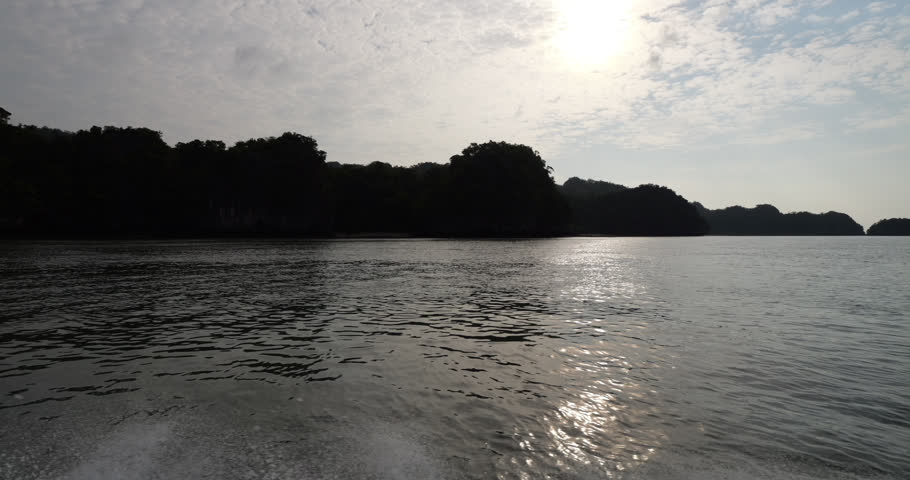 Silhouetted shoreline of Kilim Geoforest Park passes by as boat sails, with tropical trees and rugged cliffs outlined against the low bright sun. Sunlight shimmers across the calm rippling water