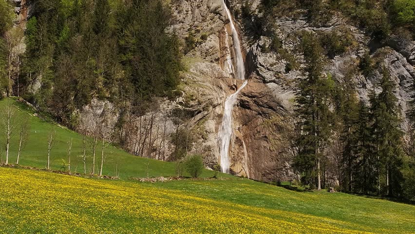 View of Sulzbachfall waterfall in Glarus, Switzerland. Scenic cascade flowing down rocky cliffs with green meadow and yellow wildflowers in the foreground, peaceful alpine nature.