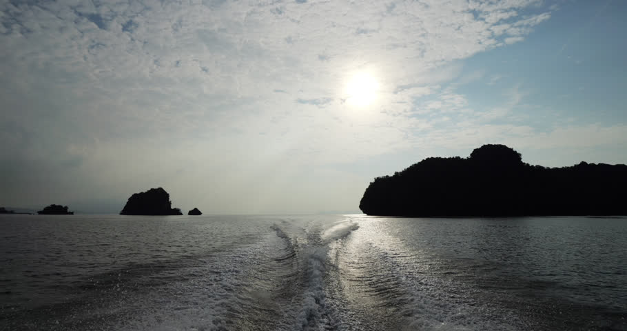 Looking back from a motorboat sailing at sunset, wake trailing across calm sea water. Silhouetted karst islets of northern Langkawi outlined against the glowing sun and partly cloudy evening sky.