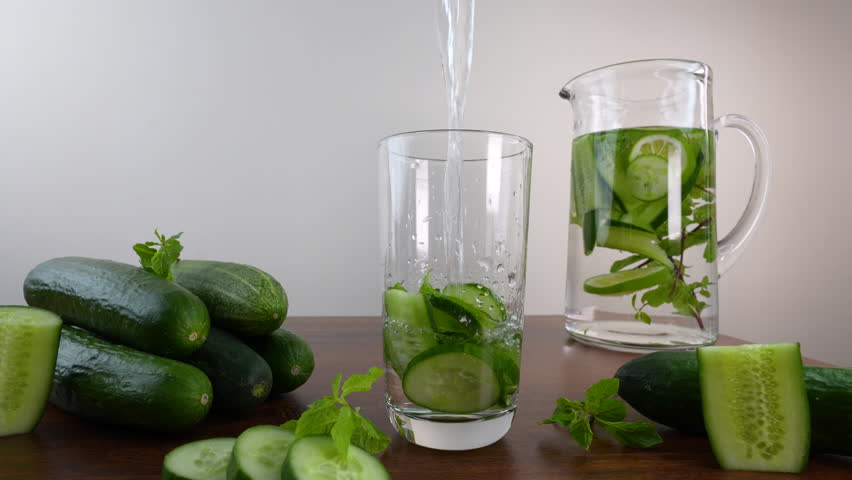 A Chilled cucumber water poured into a glass with mint leaves, surrounded by fresh cucumbers