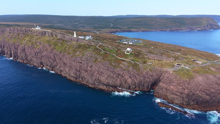 A long-range aerial of Cape Spear shows the windswept peninsula with grassy headland and swirling Atlantic waters, Signal Hill visible across the bay. Filmed from outside the national park boundary.