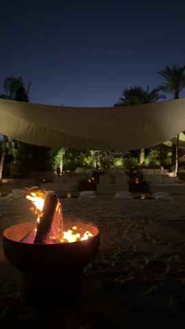 A night scene with a campfire, people sitting around, enjoying warmth and conversation. Tents and outdoor items visible, creating a cozy atmosphere. Dark sky, trees, and nature elements in the backgro