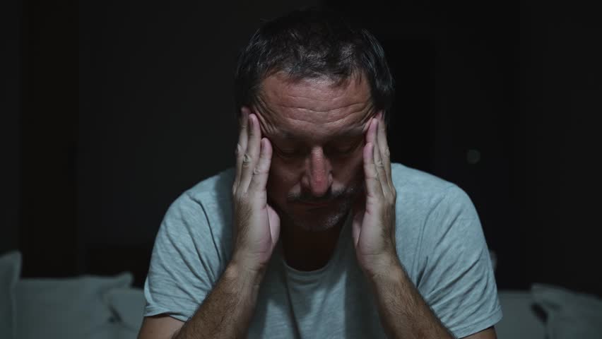 Fatigued man with closed eyes resting in dimly lit living room, symbolizing burnout and insomnia. 4K with selective focus.