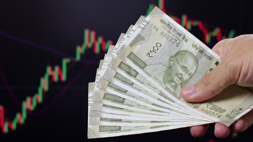 Close up view of man hands, Indian rupee banknotes, market chart on computer screen in background. Concept of currency growth, stock price change, volatile financial market, internet trading in India.