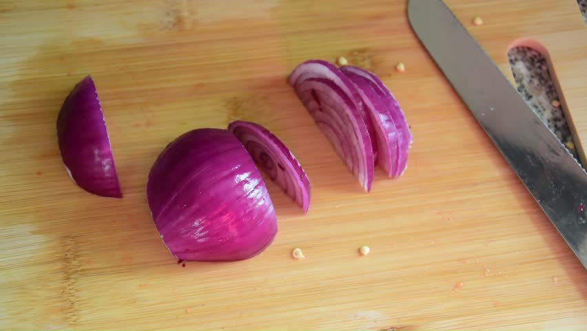 Well-lit, slow motion, close shot of someone chopping red onions on a wooden chopping board