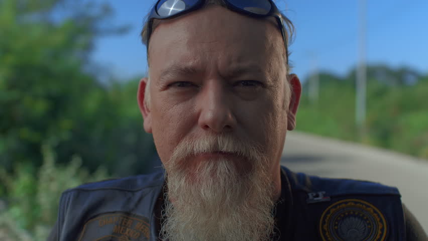 Close-up portrait of a senior man with a long white beard, helmet, sunglasses, and leather vest on a road trip. Biker lifestyle.