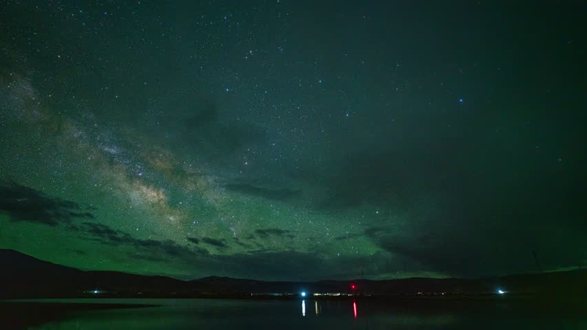 A stunning night scape featuring the Milky Way stretching across a star-filled sky above a tranquil lake. The scene is enhanced by faint light reflections on the water, adding depth and a sense