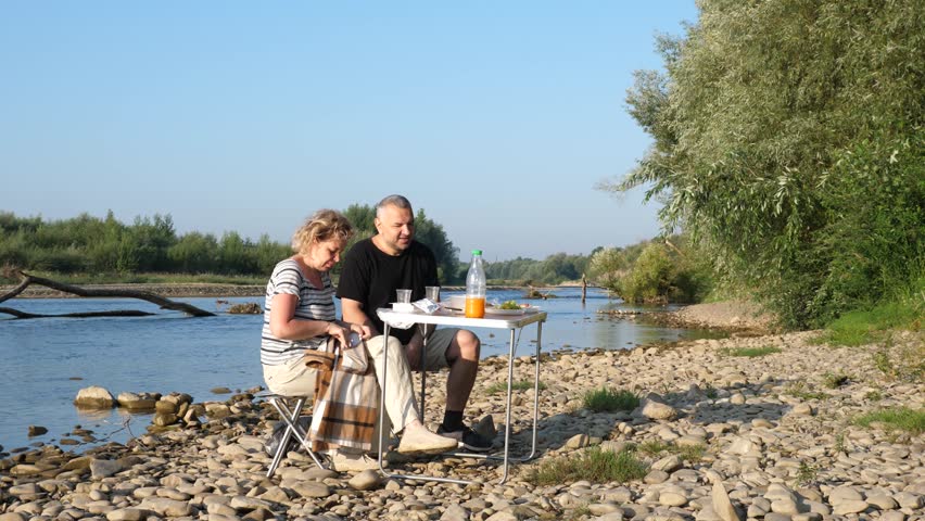 A man and woman had a picnic on the riverbank, enjoying food and drinks.
