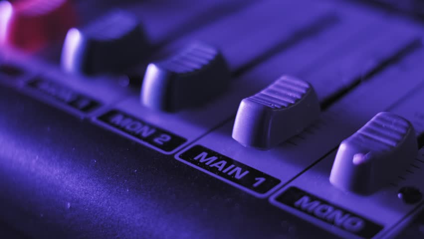 Macro shot of a finger pushing the main volume fader on an audio mixing console, showing channel labels like MAIN 1 and MON 2 under dynamic, shifting purple and orange club lights in a dark studio.