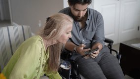 A young man in a wheelchair and his mature mother share joyful moments and photos on a smartphone in a cozy living room filled with love and connection. - Powered by Shutterstock - Get 15% off with code: PIKWIZARD15