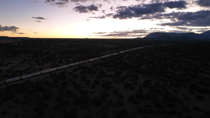 Aerial view of a train winding through the dark, arid landscape at sunset, casting long shadows across the terrain, Flagstaff, Arizona, United States.