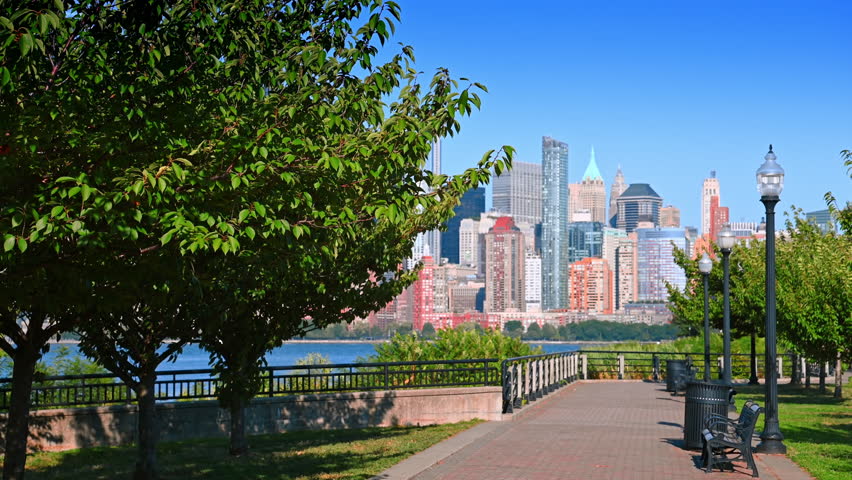Park promenade with view of Jersey City and Manhattan. A quiet park promenade in Jersey City with benches and lamps overlooking Manhattan’s skyline.